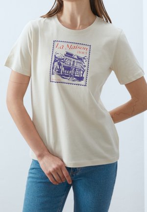 Kobieta ubrana w beżowy T-shirt z fioletowo-czerwonym motywem hotelu i napisem „La Maison Hotel” z przodu, w połączeniu z niebieskimi dżinsami.