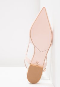 Rosa klackskor med spetsig tå och slät yta, genomskinliga remmar och texturerad sula. Har en gulddetalj nära bakdelen.