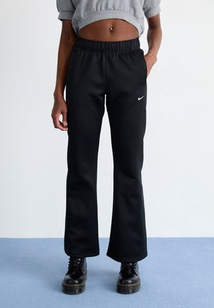 Person iført sorte Nike flared bukser, cropped grå sweatshirt, sorte platform snørestøvler, stående på blå gulv med hvid baggrund.