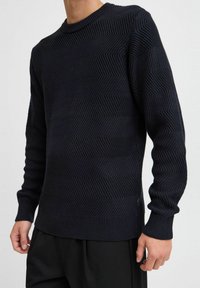 Marineblå strikkegenser med et teksturert sikksakkmønster, rund hals og ribbestrikkede mansjetter og kant. Tettsittende design med lange ermer.