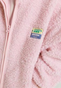 Chaqueta de forro polar rosa con una superficie suave y texturizada y una cremallera a juego. Presenta una etiqueta colorida que dice "LA MAISON AMV SUPER VINTAGE".