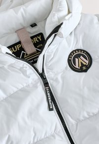 Giacca a vento bianca con cappuccio, texture trapuntata, zip frontale e patch logo nero e oro sul petto, con una linguetta beige all'interno.