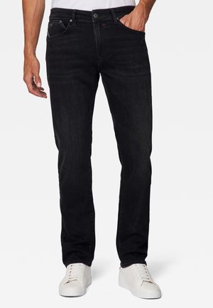 Uomo che indossa jeans neri dritti e sneakers bianche, in piedi con una mano in tasca su sfondo uniforme.