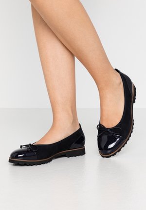 Ben klædt i sort ruskind og patenteret læder balletsko med sløjfedetalje, stående på et glat hvidt gulv.