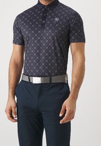 Marinblå polo-shirt med geometriskt mönster, korta ärmar och en tvåknapplåst. Matchas med grått bälte och mörka byxor.