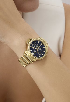 Reloj de pulsera dorado con esfera negra, números romanos, tres subesferas y un bisel adornado con cristales en la muñeca de una persona.