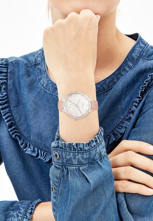 Zilverkleurig horloge met een lichtroze siliconen band, ronde wijzerplaat met decoratieve elementen en duidelijke uurmarkeringen, gedragen op een denim blouse.