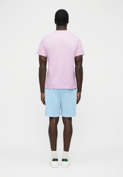 Άνδρας που στέκεται γυρισμένος προς τα πίσω, φοράει ροζ λευκό κοντομάνικο πουκάμισο, ανοιχτό μπλε σορτς, λευκές κάλτσες και λευκά αθλητικά παπούτσια μπροστά σε απλό φόντο.