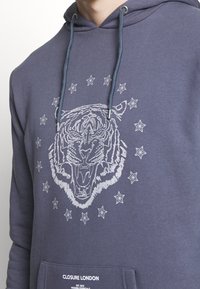 Felpa con cappuccio in tessuto blu navy, caratterizzata da una grafica di una tigre bianca circondata da stelle. Include una tasca frontale e un cordoncino.