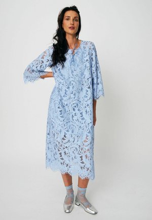 Woman with long dark hair wearing a light blue lace midi dress with three-quarter sleeves and silver shoes with sheer patterned socks.