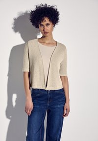 Beige Strickjacke mit kurzen Ärmeln, strukturiertem Gestrick und offener Front, getragen über einem hellen Oberteil und kombiniert mit dunkelblauen Jeans.