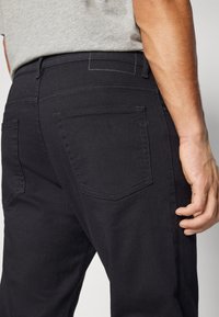 Jean en denim noir avec une coupe droite, poches arrière avec des coutures discrètes et une texture lisse. Une chemise gris neutre est partiellement visible.