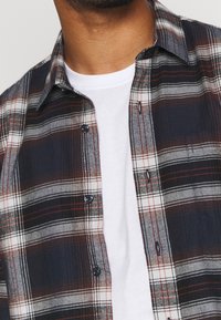 Koszula w kratę w kolorze granatowym, białym i bordowym, wykonana z materiału o miękkiej teksturze, z guzikami i kołnierzykiem. Nosi się ją na białej koszulce.