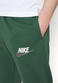 Person som bär gröna Nike träningsbyxor med handen i sidofickan och vit t-shirt synlig vid midjan.