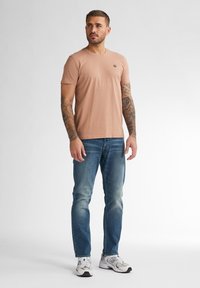 T-shirt rosa chiaro a maniche corte con scollatura rotonda e piccolo logo, abbinato a jeans blu di lavaggio medio e sneakers bianche.