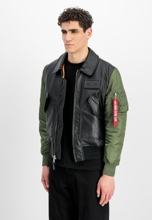 Man met een zwarte leren jas met groene stoffen mouwen en een rood ritslabel op de linkerarm, staand tegen een effen witte achtergrond.