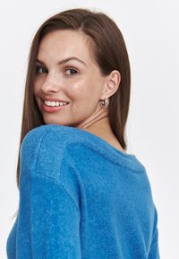 Niebieski sweter z dzianiny o luźnym kroju, okrągłym dekolcie i teksturowanej tkaninie. Modelka nosi srebrne kolczyki w kształcie kół, zwrócona nieco w tył.