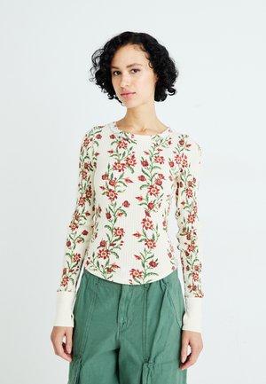Langarmtop met een geribbelde textuur, witte basis, bloemmotief in rood en groen, en contrasterende crèmekleurige manchetten bij de polsen.