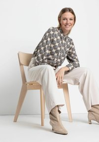Langärmlige Bluse mit schwarzem und beige geometrischem Muster, kombiniert mit lockeren cremefarbenen Hosen und tan-farbenen Wildleder-Stiefeletten, sitzend auf einem Holzstuhl.