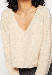 Pull beige surdimensionné en maille avec un col en V profond, une texture côtelée et des épaules tombantes, associé à un pantalon sombre taille haute.