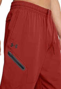Rote Sporthose mit elastischem Bund und Kordelzug, ausgestattet mit einer seitlichen Reißverschlusstasche und schwarzen Akzenten. Leichtes Material und schmal zulaufendes Design.