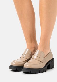 Mocassins en cuir verni beige avec une plateforme noire épaisse, présentant une pointe incurvée et deux bandes croisées à l'avant.