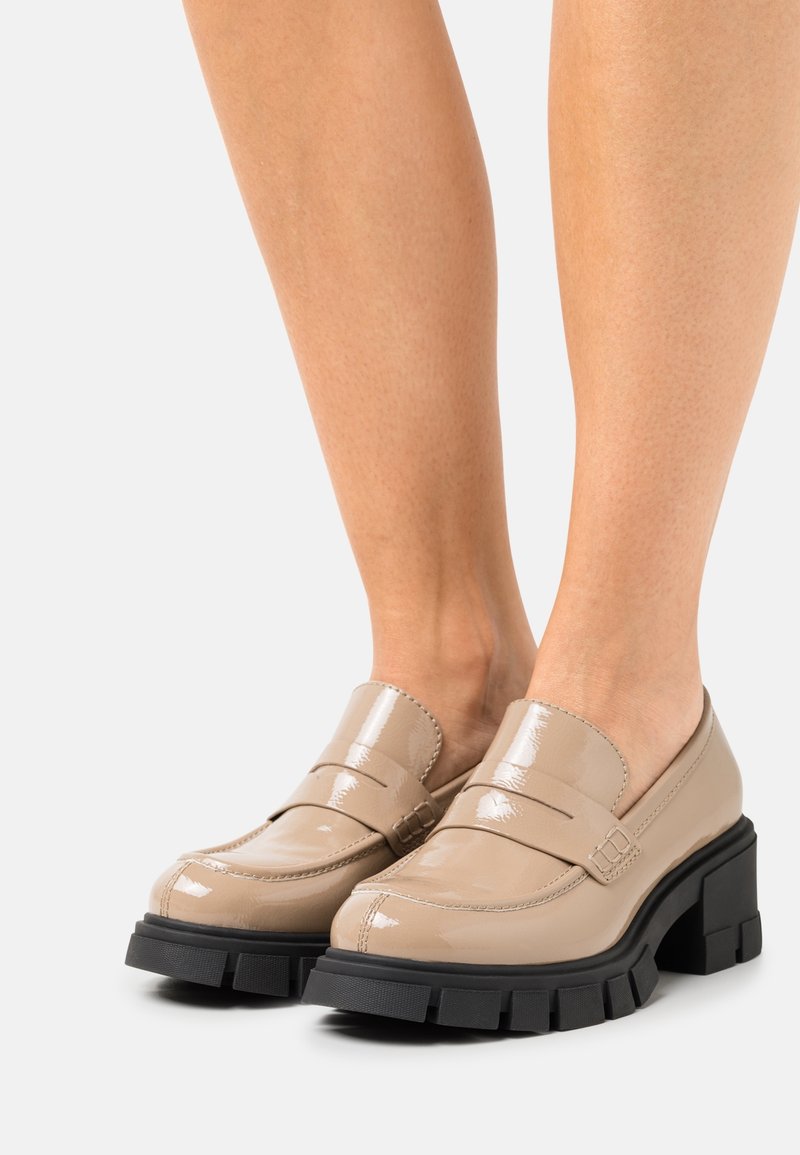 Mocassins en cuir verni beige avec une plateforme noire épaisse, présentant une pointe incurvée et deux bandes croisées à l'avant.
