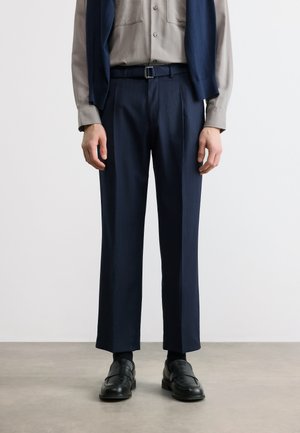 Άνδρας που φοράει σκούρο μπλε παντελόνι καλοραμμένο, μαύρη ζώνη, μαύρα δερμάτινα μοκασίνια, γκρι πουκάμισο με κουμπιά και σκούρο μπλε πουλόβερ περασμένο πάνω από τους ώμους.