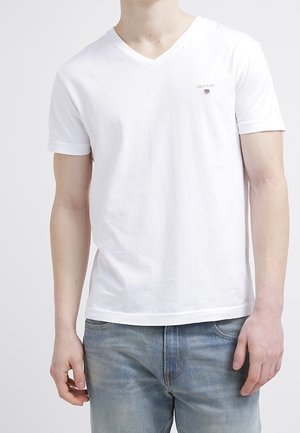 Mężczyzna w gładkiej białej koszulce z dekoltem w serek i małym logo GANT na lewej piersi oraz w jasnoniebieskich dżinsach, stojący na białym tle.