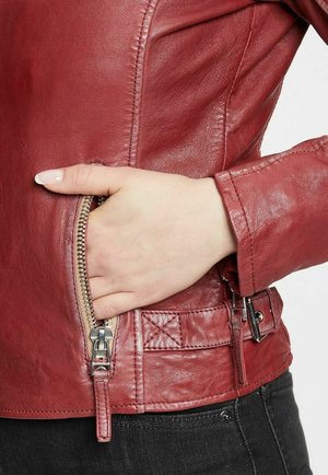 Veste en cuir rouge avec un gros plan d'une main dans une poche zippée sur le côté. Présente une texture lisse et un matériel de fermeture éclair métallique.