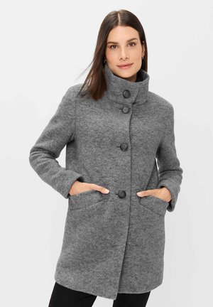 Frau mit glatten braunen Haaren, die einen grauen knöpfbaren Mantel mit hohem Kragen trägt und die Hände in den vorderen Taschen hat, vor einem weißen Hintergrund stehend.