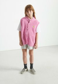 Roza pletena brezrokavnik z rebrasto teksturo, oversize kroj in gumbnim zapenjanjem. Nosi se čez belo majico in v kombinaciji s sivimi kratkimi hlačami ter karo čevlji.