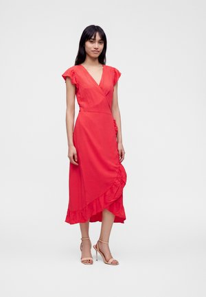 Femme debout portant une robe portefeuille rouge sans manches avec des détails à volants et des sandales à talons beiges sur un fond blanc uni.