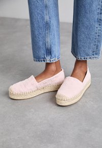 Espadrilles de color rosa claro hechas de tela acolchada con una suela de cuerda de yute, con punta redondeada y sin tacón. Llevadas con jeans cortos azules.