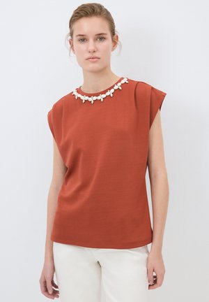 Top corto color ruggine con maniche a sbuffo, caratterizzato da un tessuto testurizzato e un accentuazione di perline bianche e fiori al colletto. Vestibilità semplice e rilassata.