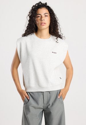 Vrouw met krullend haar, gekleed in een lichtgrijze sweatshirt zonder mouwen met opgevulde schouders en de tekst "New York", en grijze broek, met haar handen in de zakken.