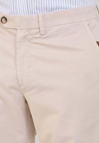 Pantaloni in cotone beige con una superficie liscia, dotati di tasche laterali e chiusura con bottone singolo in vita. Design semplice e pulito.