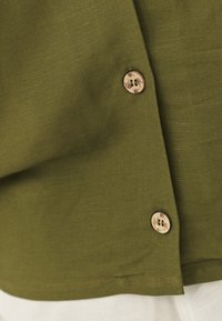 Cămașă verde măsliniu dintr-un material texturat, cu nasturi din lemn. Designul include o margine rotunjită și detalii cusute la cusături.