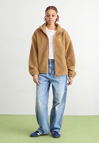 Geacă din polar bej cu fermoar și mâneci lungi, purtată peste un tricou alb. Asortată cu blugi largi albaștri și pantofi sport albaștri cu dungi albe.