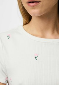 T-shirt blanc à rayures avec une petite broderie de rose rose sur les épaules. Encolure ronde et manches courtes. Texture lisse.