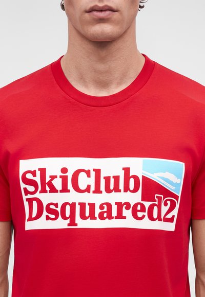 Крупен план на човек, носещ червена тениска с голямо бяло лого „SkiClub Dsquared2“ и графика на скиор.