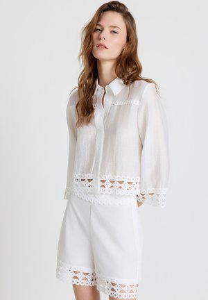 Giovane donna con capelli lunghi che indossa una camicetta bianca trasparente e pantaloncini bianchi, entrambi con bordo in pizzo sangallo, in piedi davanti a uno sfondo uniforme.