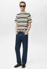 Chemise à rayures à manches courtes en crème, vert et rouge ; associée à un jean bleu foncé à coupe ample et des chaussures noires. Modèle portant des lunettes de soleil.