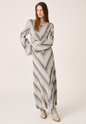 Femeie purtând o rochie lungă cu model în zigzag, mâneci largi și cizme texturate până la gleznă, stând cu brațele încrucișate și privirea în lateral.