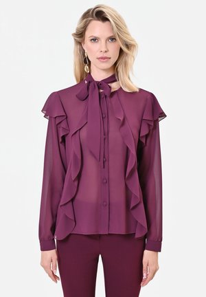 Donna che indossa una camicetta color prugna trasparente con ruches sul davanti e un fiocco legato al collo, abbinata a pantaloni coordinati color prugna su sfondo bianco.