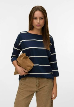 Femme aux cheveux bruns raides portant un haut rayé bleu marine et blanc et un pantalon beige, tenant une pochette en daim beige.
