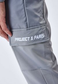 Grå stofbukser har en sidelomme med teksten "PROJECT X PARIS" i hvid broderi. Glat tekstur og skræddersyet design.