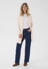 Mujer de pie vestida con un cárdigan beige claro, camisa blanca, vaqueros oscuros y botas de tobillo marrones frente a un fondo blanco liso.