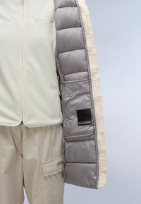 Chaqueta de forro polar color beige claro con un interior acolchado gris, cierre de cremallera y un forro texturado color crema. Presenta un parche de logo en el interior.