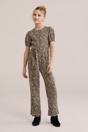 Mlada djevojka stoji noseći kombinezon u boji preplanule i crne leopard uzorka s puf rukavima i crnim gležnjačama.